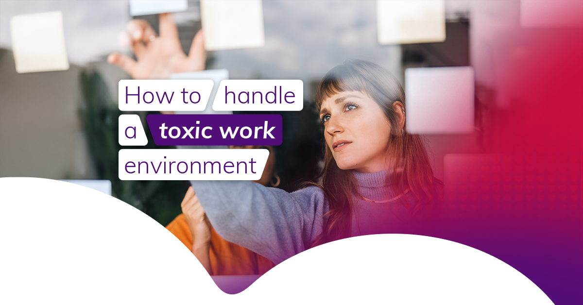 A brunette woman wearing a grey sweater is at work and pointing at something on a board. It appears she's in a workshop with others