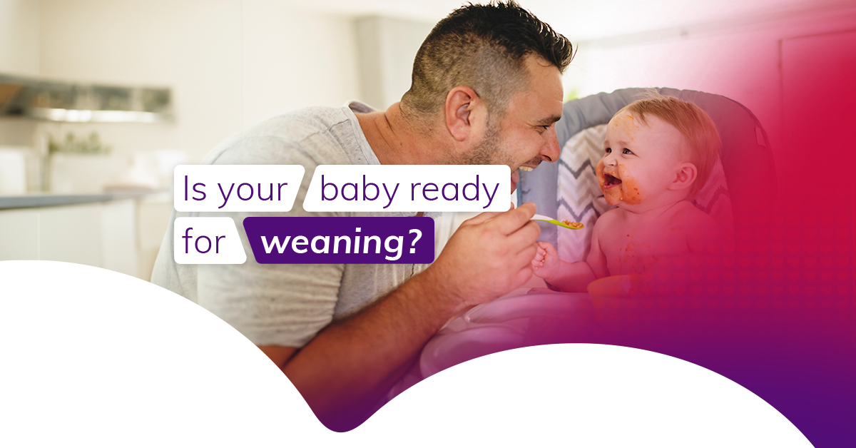 A father spoon feeds his smiling baby who is sitting in a baby chair